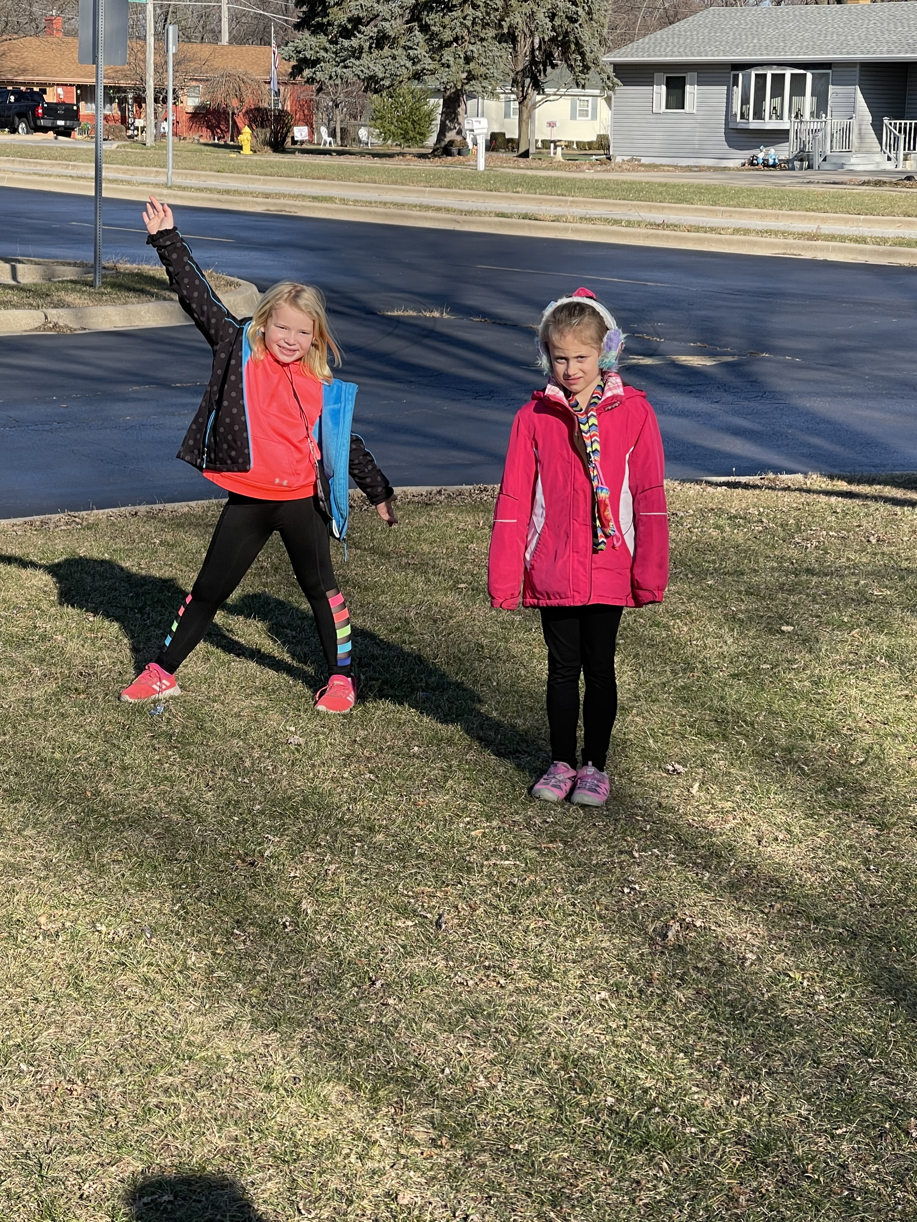 Two young girls are standing on a grassy area, one is waving enthusiastically while the other stands beside her, both dressed in colorful jackets and athletic wear, with a suburban street visible in the background.