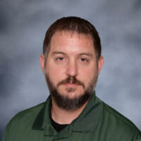 A man with a beard wearing a green shirt, standing confidently with a neutral expression.