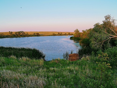 A serene lake bordered by lush grass and trees, illuminated by the warm hues of a sunset.