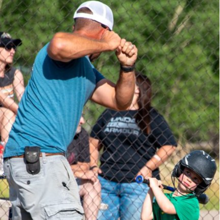 A man guides a young boy as he swings a bat at a ball in an outdoor setting, showcasing a moment of learning and play.