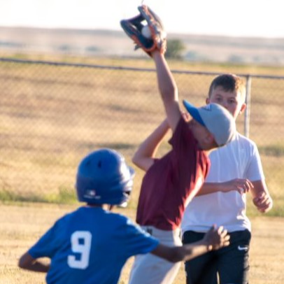 A young boy stretches his arm upward, eagerly reaching for a baseball in a bright outdoor setting.