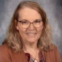 A woman wearing glasses and a brown cardigan, smiling softly while looking towards the camera.