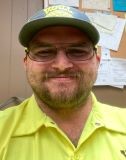 A man wearing a yellow shirt and hat smiles warmly, exuding a cheerful and friendly demeanor.