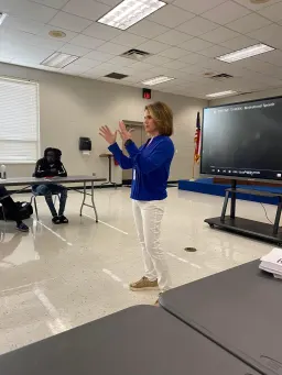 A woman stands in front of a classroom, addressing an audience seated at tables, engaged in learning.