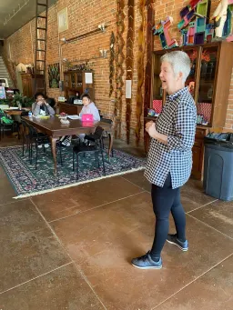 A woman stands in a room while people are seated at tables, engaged in conversation and activities.