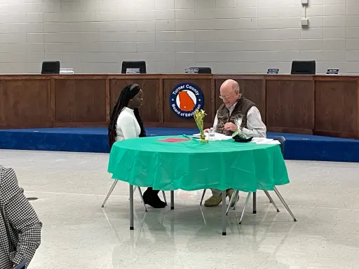 A man and woman seated at a table, engaged in conversation within a cozy room setting.