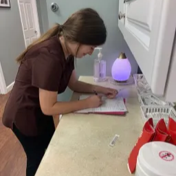 A woman is focused on writing on a piece of paper while seated at a kitchen table.