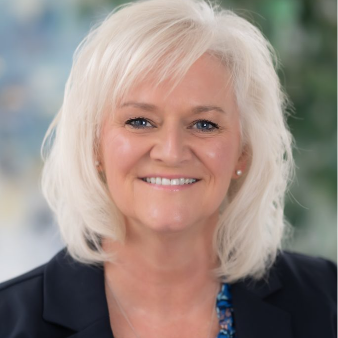 A smiling woman with shoulder-length blonde hair, wearing a black blazer over a blue floral blouse, stands in front of a blurred background with greenery.
