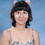A person with short, dark hair and pink highlights is wearing a floral dress and pink cat ears, posing against a blue background.