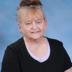 A woman with light brown hair styled in a bun, wearing a black shirt over a purple top, poses against a light blue background.