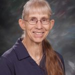 A smiling woman with long, light-colored hair and glasses, wearing a dark shirt, poses for a portrait against a blurred background.