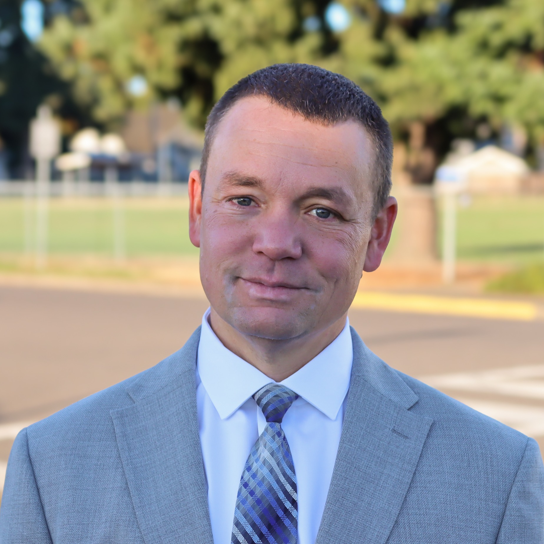 A smiling man with short hair wearing a black polo shirt with a logo, set against a street background.