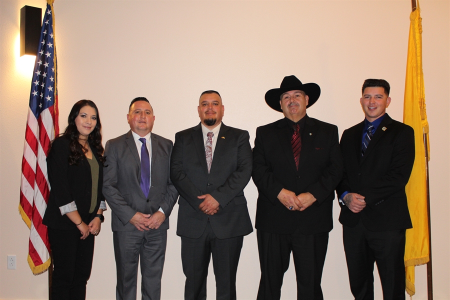A group of five individuals, including a woman and four men in formal attire, stand together in front of the American and New Mexico flags.
