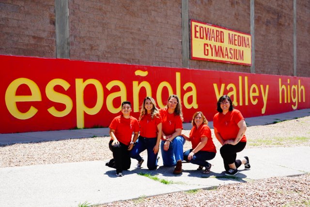 A group of six women in red shirts poses in front of a large wall sign that reads 'española valley high' and 'EDWARD MEDINA GYMNASIUM' in yellow letters.