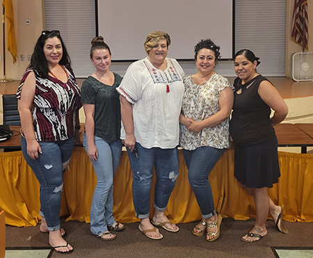 Five women stand together in a room, smiling and posing for the camera, with a projector screen in the background and a table in front of them.
