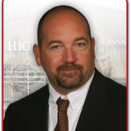 A man with a shaved head and a goatee, wearing a black suit and a white shirt with a brown tie, stands in front of a blurred background that suggests a school or educational institution.