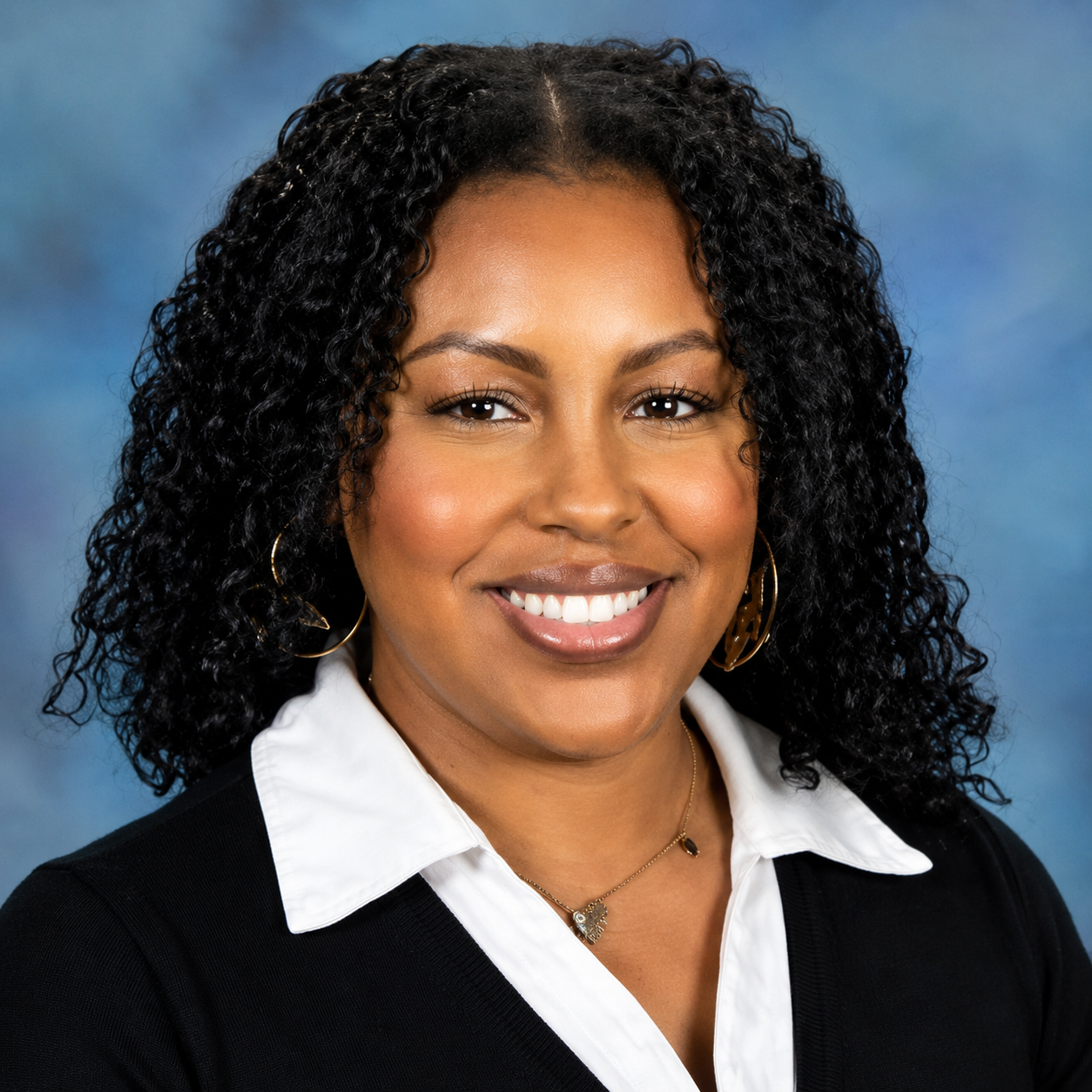 Professional headshot of a smiling woman with shoulder-length curly dark hair, wearing a white collared shirt under a black sweater. She is facing the camera against a soft blue studio background, with gold hoop earrings and a delicate necklace. The portrait conveys a warm, confident, and approachable professional appearance.