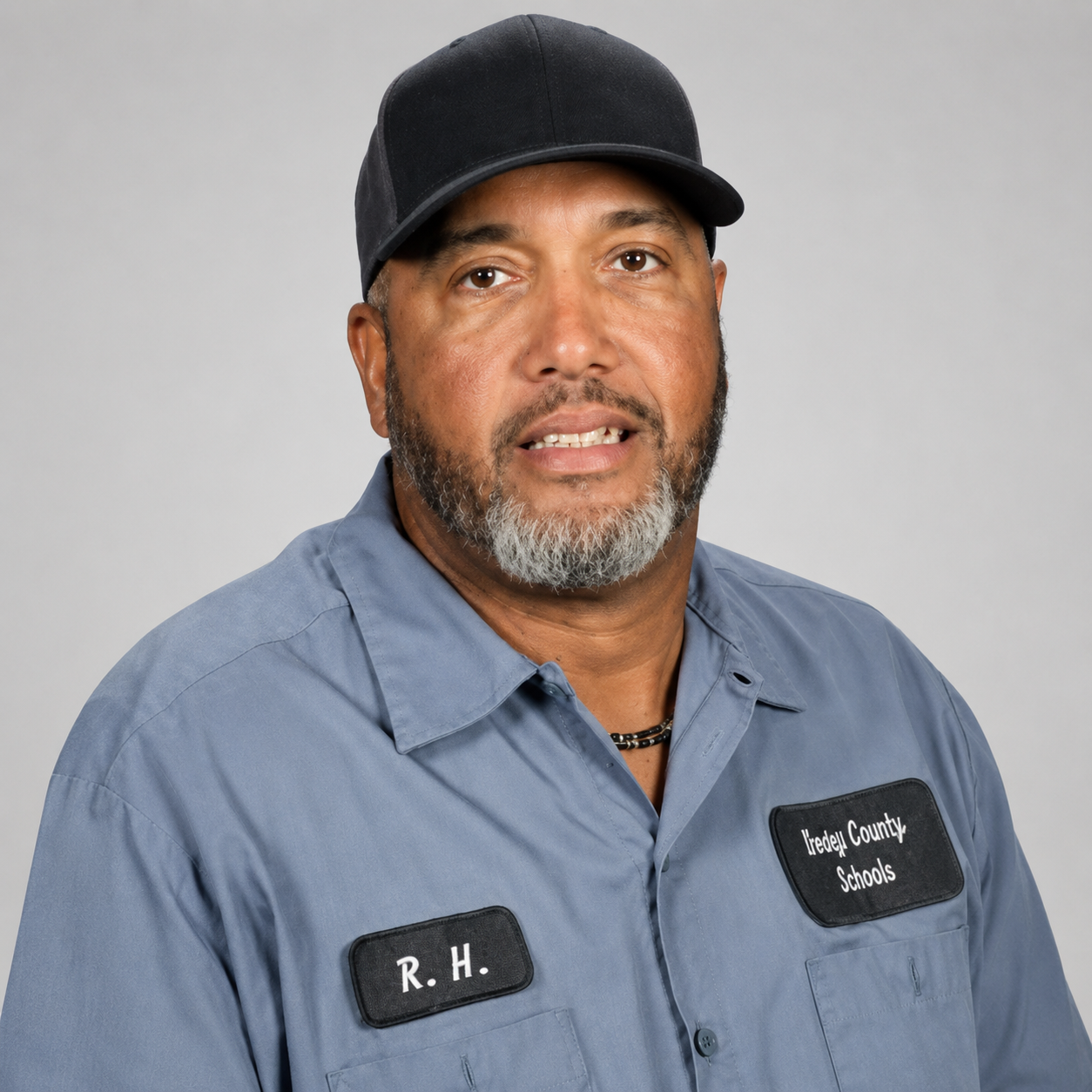 Professional headshot of a man wearing a black cap and blue work shirt against a light gray background.