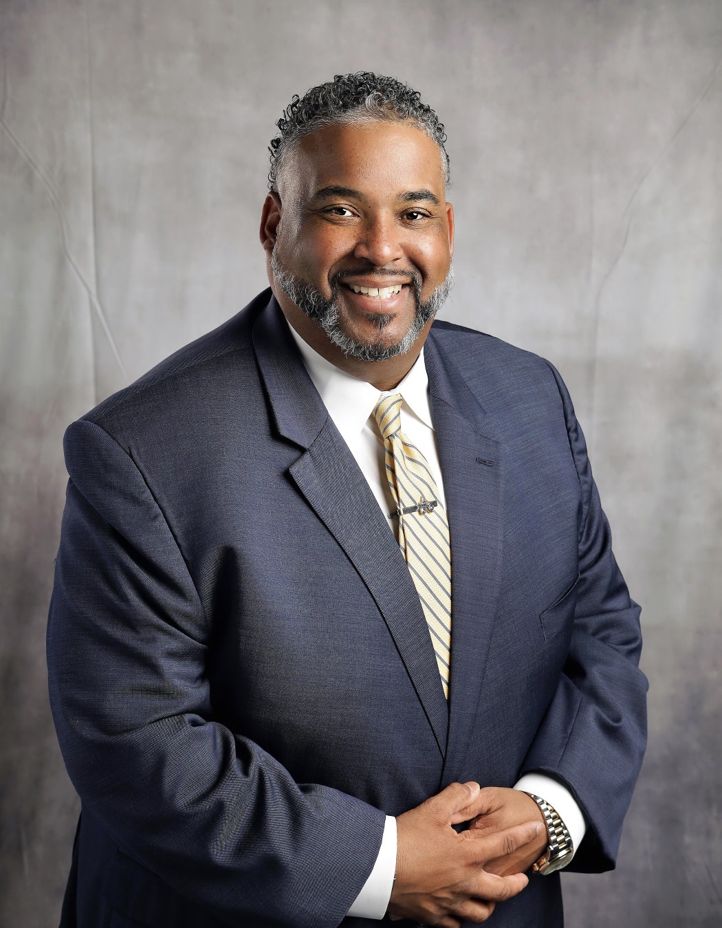 Dr. Giron Wooden: Professional portrait of a smiling man wearing a navy suit, white shirt, and gold-and-blue striped tie.