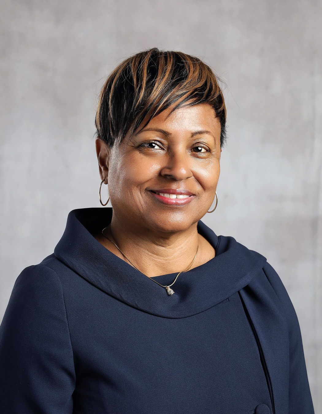 Melissa Harvey: Professional portrait of a smiling woman wearing a navy dress, hoop earrings, and a gold necklace.