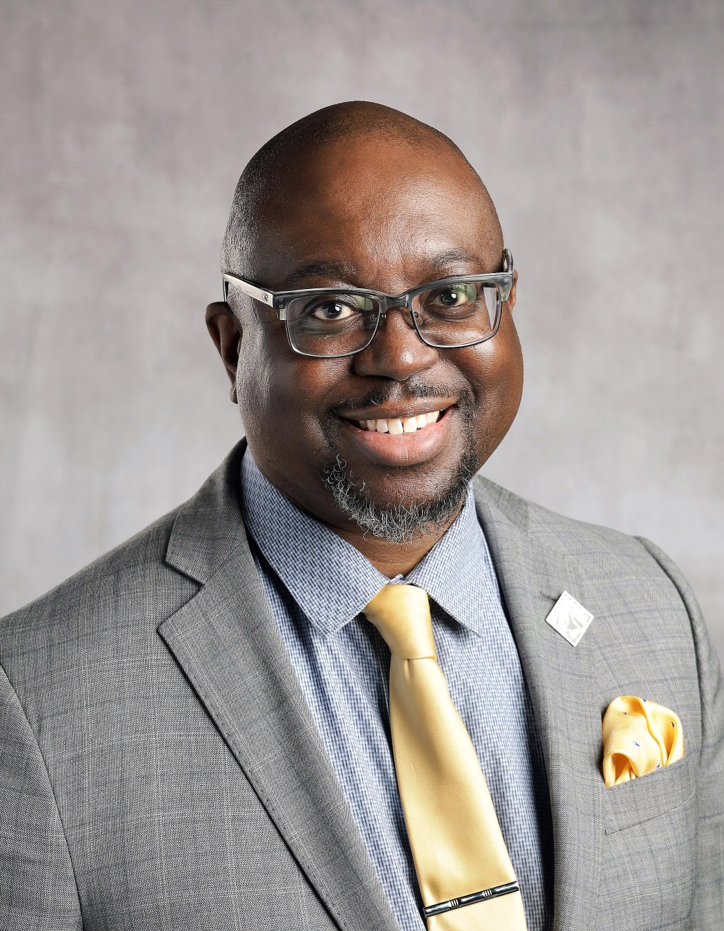 Preston Mckellar: Professional portrait of a smiling man wearing glasses, a gray suit, blue shirt, and gold tie.