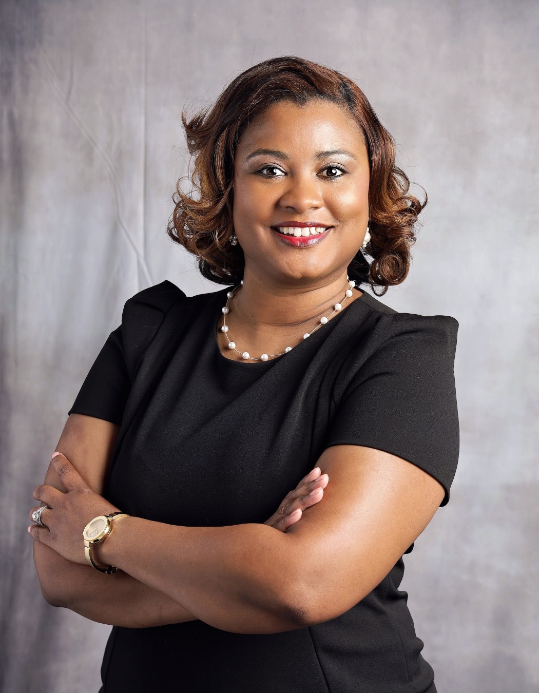 Brittney Custalow: Professional portrait of a smiling woman wearing a black dress and pearl necklace with her arms crossed.
