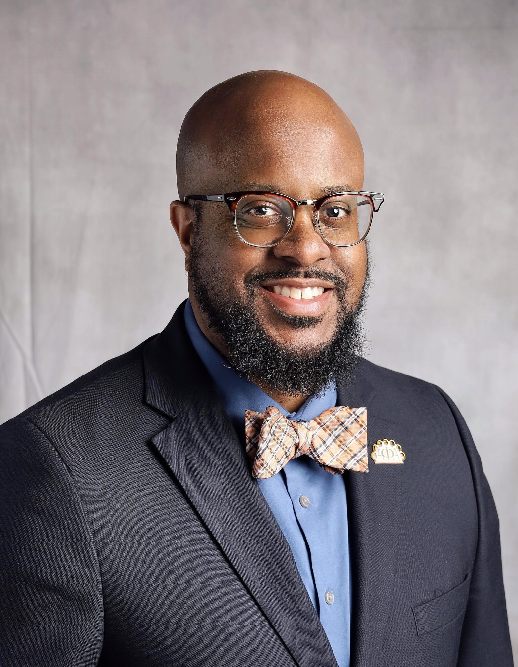 Patrick King: Professional portrait of a smiling man wearing glasses, a navy suit, blue shirt, and plaid bow tie.
