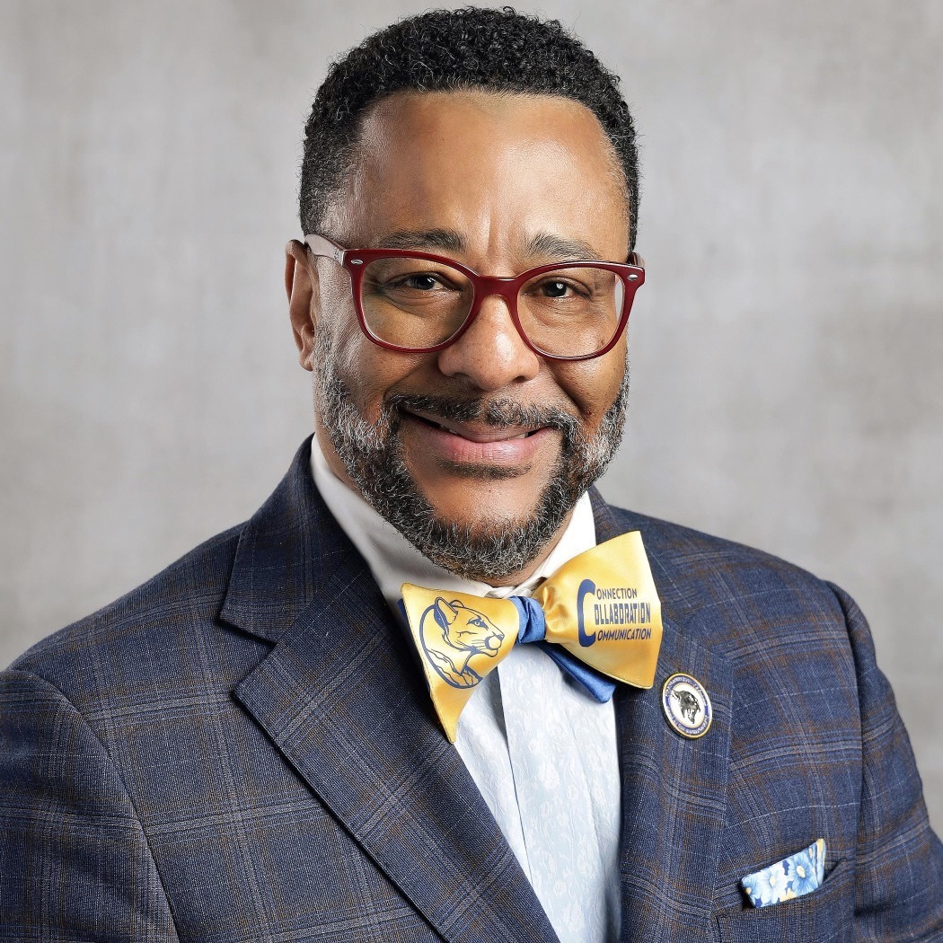 Professional headshot of a smiling male school district administrator wearing red-framed glasses, a blue plaid suit jacket, white dress shirt, and a gold-and-blue bow tie featuring a cougar logo and the words "Connection, Collaboration, Communication." He is posed against a light gray studio background and wears a circular lapel pin and patterned pocket square.