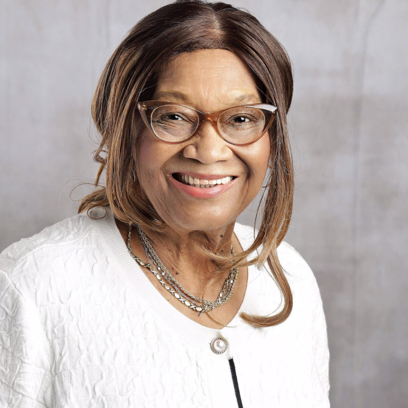 Smiling woman wearing glasses, a white sweater, and layered silver necklaces against a light gray background.