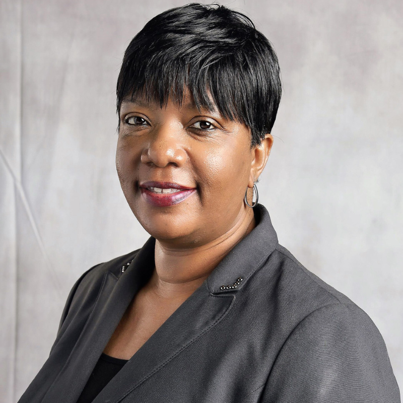 Woman with short black hair wearing a dark blazer and hoop earrings, smiling against a light gray background.