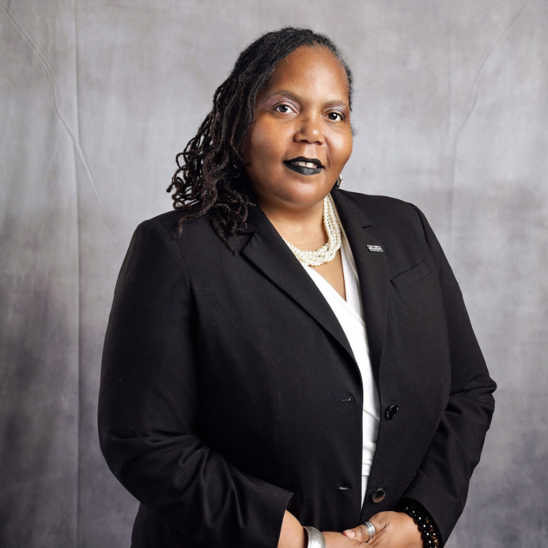 Woman with locs wearing a black blazer, white top, and pearl necklace, posed against a gray background.