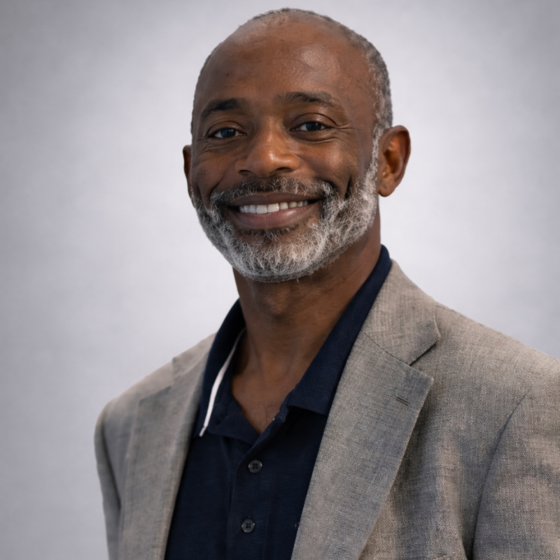 Smiling man with a gray beard wearing a light gray blazer and navy polo shirt against a light gray background.