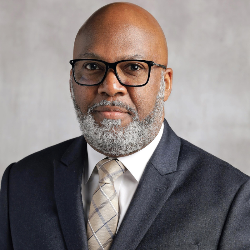 Man with glasses and a gray beard wearing a dark suit, white shirt, and patterned tie against a light gray background.