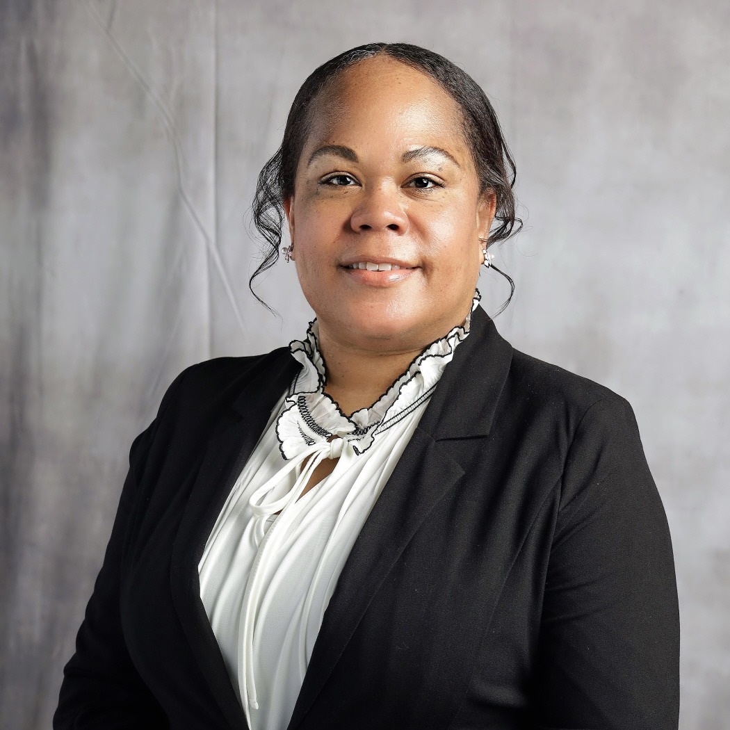 Woman wearing a black blazer and white ruffled blouse, smiling against a light gray background.