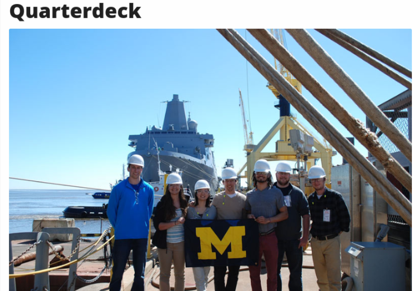 A group of six individuals wearing hard hats stands on a ship's deck, holding a flag with the letter 'M', with a large vessel in the background under clear blue skies.