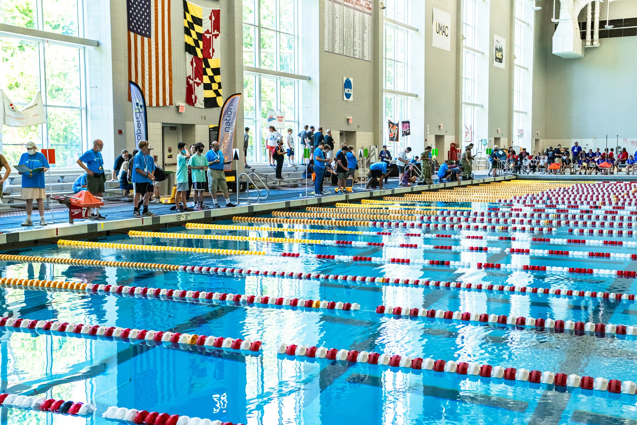 A spacious indoor swimming pool with multiple lanes, featuring swimmers and coaches preparing for a competition, and large windows allowing natural light to fill the area.