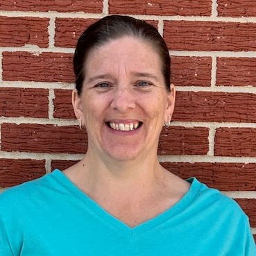  A woman with long hair wearing a gray shirt, standing confidently with a neutral expression.