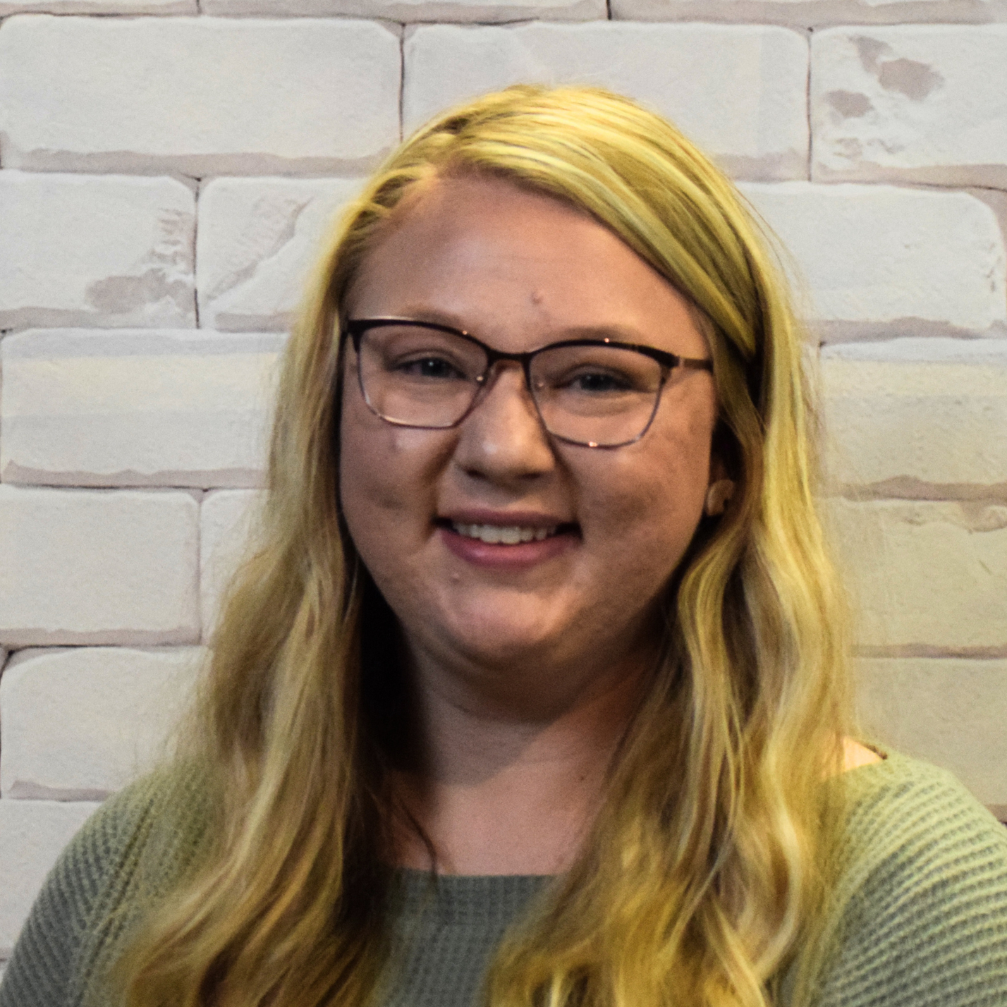 A woman wearing glasses and a brown shirt smiles warmly at the camera, exuding a friendly and approachable demeanor.