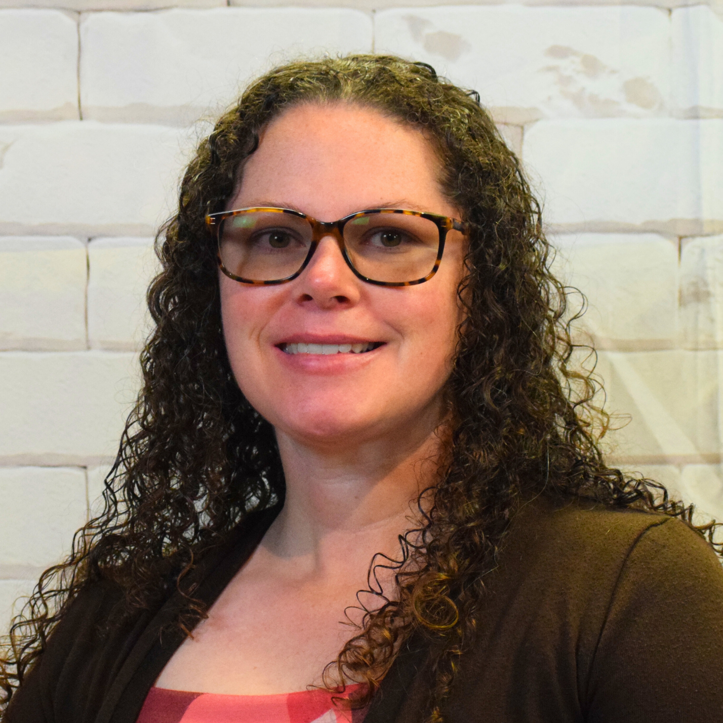 A woman with curly hair and glasses smiles warmly, exuding joy and confidence in her expression.