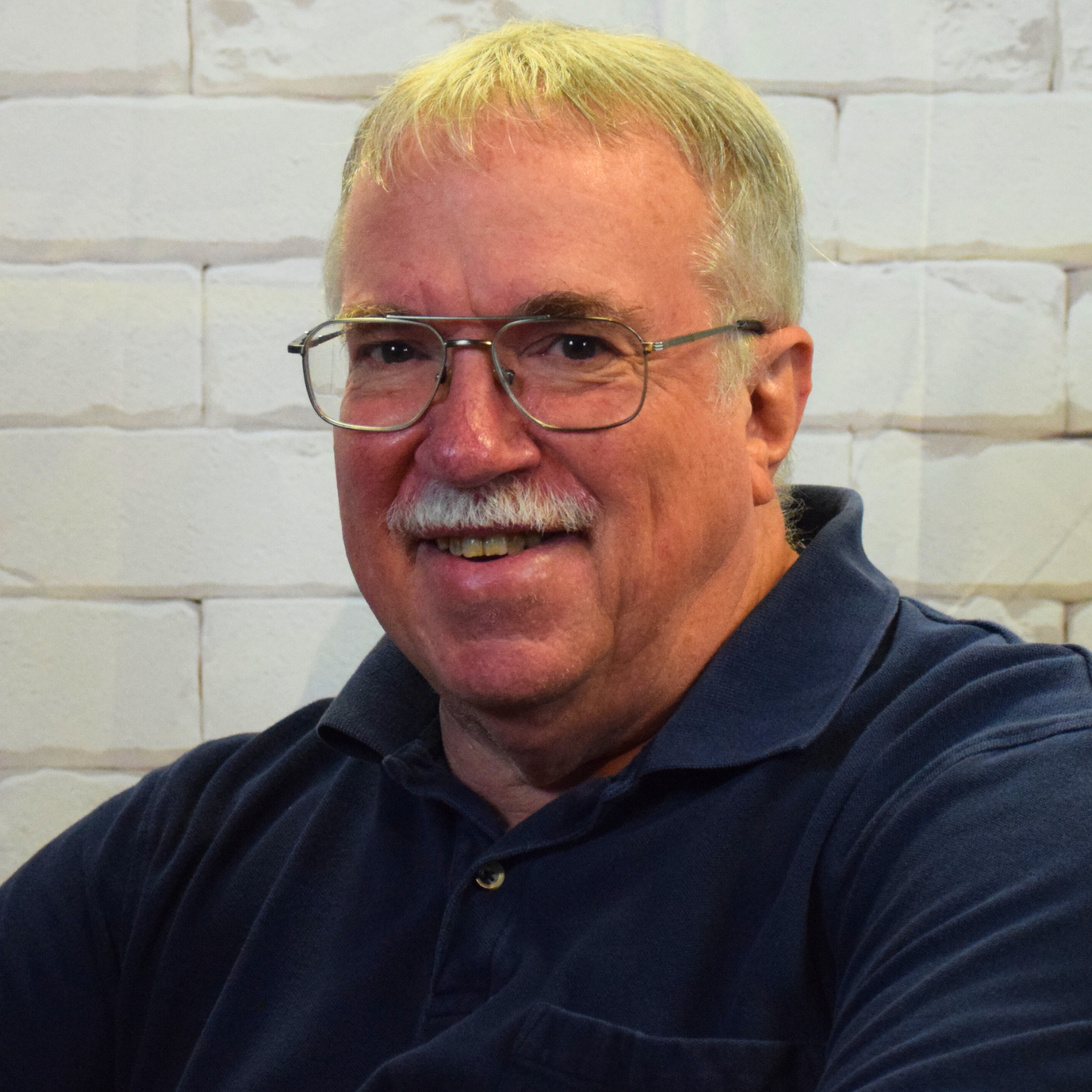  A man with glasses and a mustache sits thoughtfully in front of a textured brick wall.