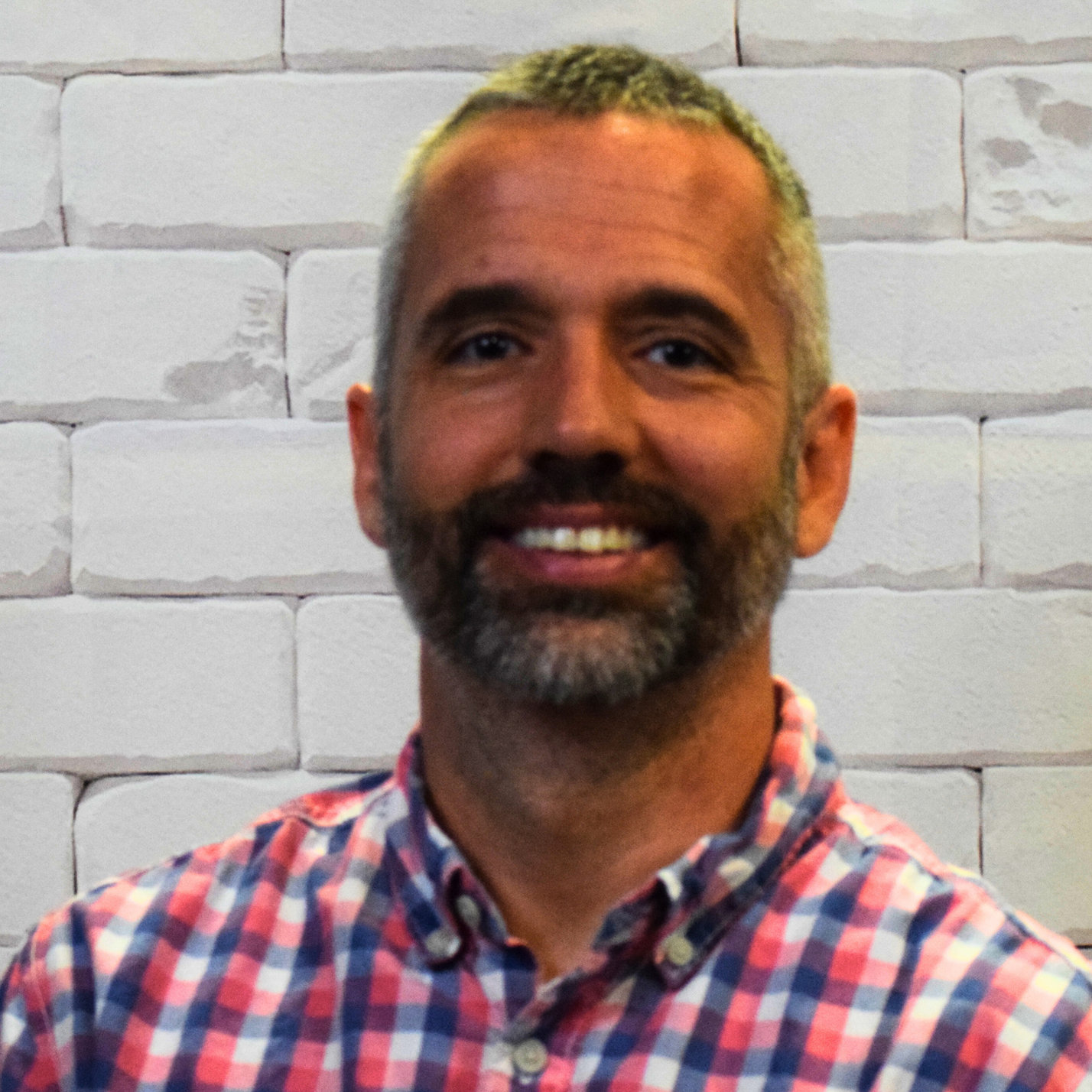  A man wearing a blue and white checked shirt smiles warmly at the camera, exuding a friendly demeanor.