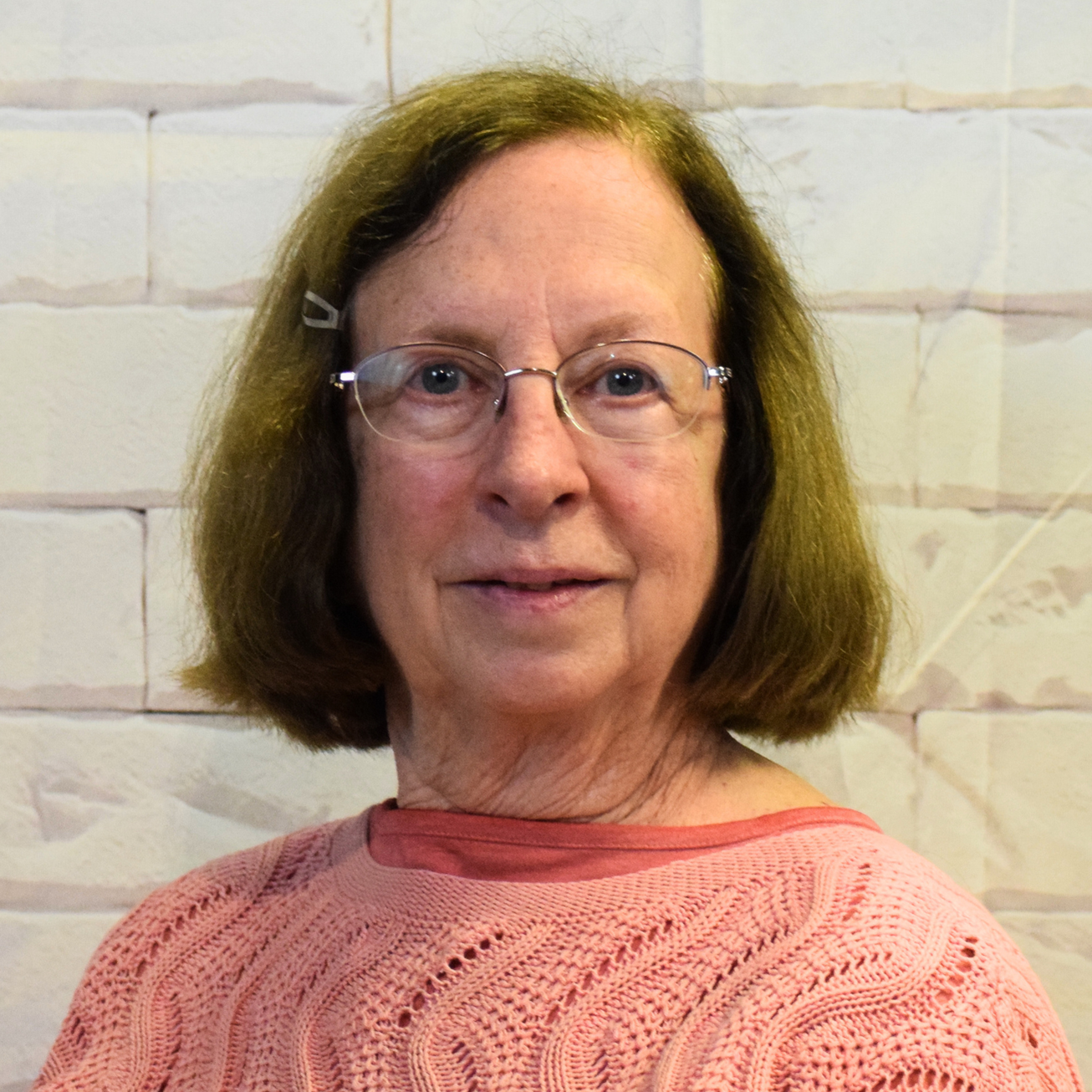 An older woman wearing glasses and a blue sweater, smiling gently while seated in a cozy environment.