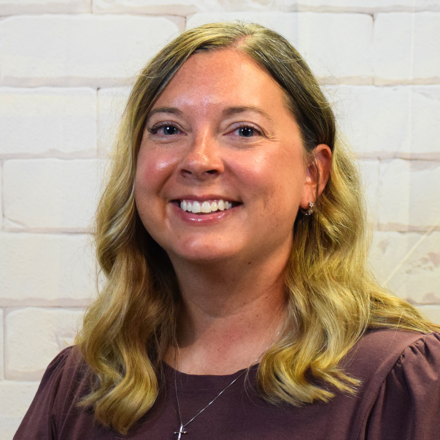 A woman with blonde hair wearing a floral shirt, smiling and standing against a neutral background.