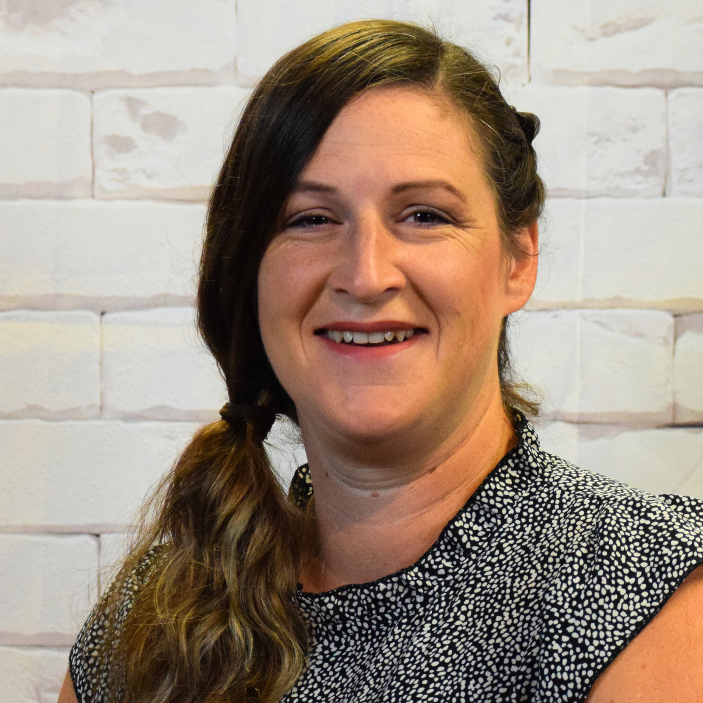 A woman wearing a blue shirt and vest stands confidently in front of a plain white wall.