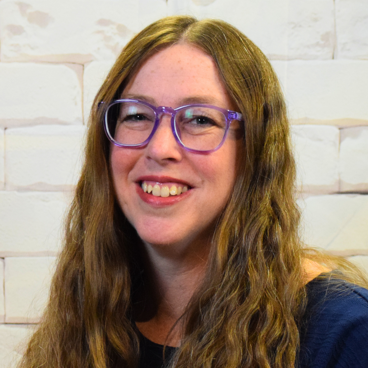 A woman wearing glasses and a pink shirt smiles warmly, exuding a cheerful and friendly demeanor.