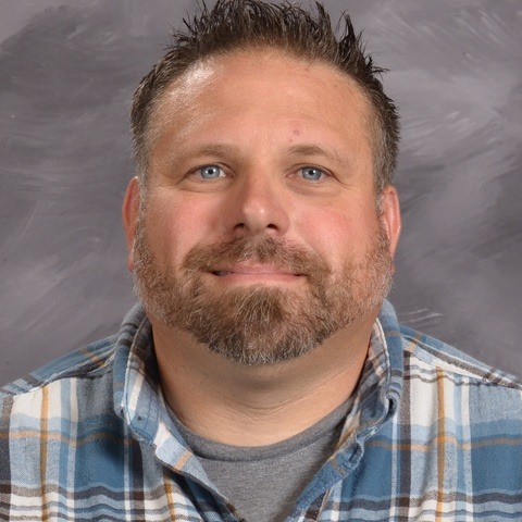 A man wearing a blue shirt smiles warmly at the camera, exuding a friendly and approachable demeanor.