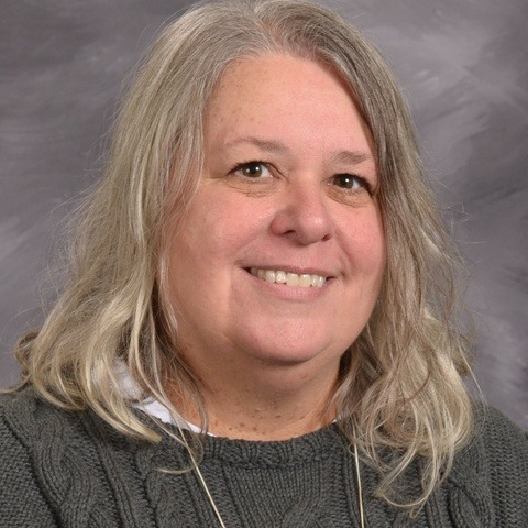 A woman with blonde hair wearing a blue shirt, smiling and standing against a neutral background.