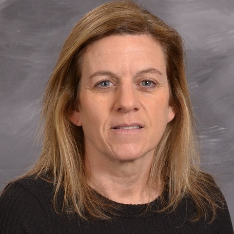 A woman wearing a green jacket over a floral shirt, standing confidently with a pleasant expression.