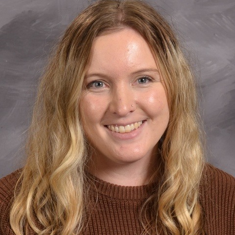 A woman with long blonde hair is wearing a blue and white striped shirt, standing against a neutral background.