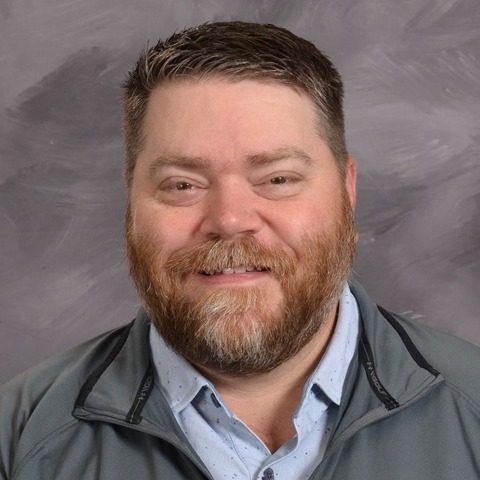 A man with a beard wearing a blue shirt, standing confidently with a neutral expression.